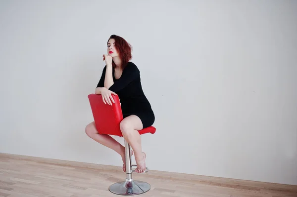 Rudowłosy dziewczyna na czarna sukienka tunika siedzi na przeciw czerwone krzesło — Zdjęcie stockowe