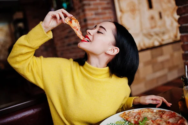 Menina morena engraçada em suéter amarelo comendo pizza no restaurante — Fotografia de Stock