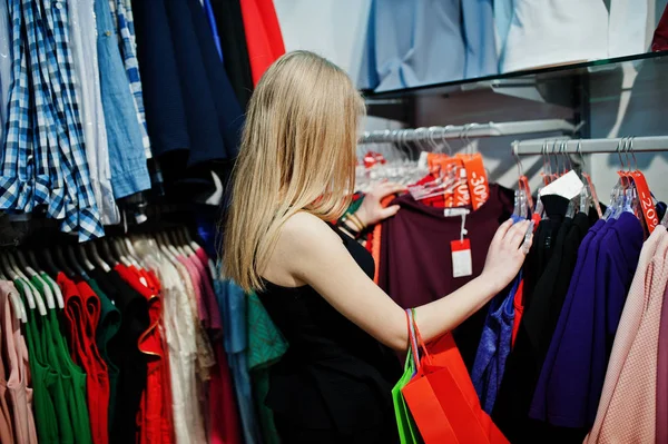 Ragazza bionda con borse della spesa sceglie i vestiti nella boutique . — Foto Stock
