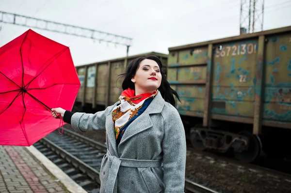 Chica morena en abrigo gris con paraguas rojo en la estación de tren . — Foto de Stock