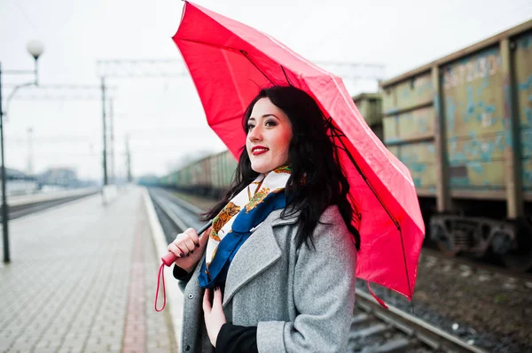 Brünettes Mädchen im grauen Mantel mit rotem Regenschirm im Bahnhof. — Stockfoto