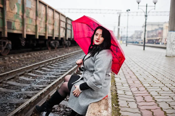 Брюнетка в сером пальто с красным зонтиком на вокзале . — стоковое фото
