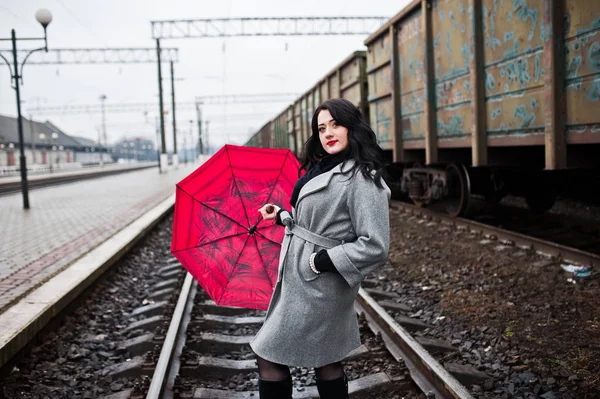 Брюнетка в сером пальто с красным зонтиком на вокзале . — стоковое фото