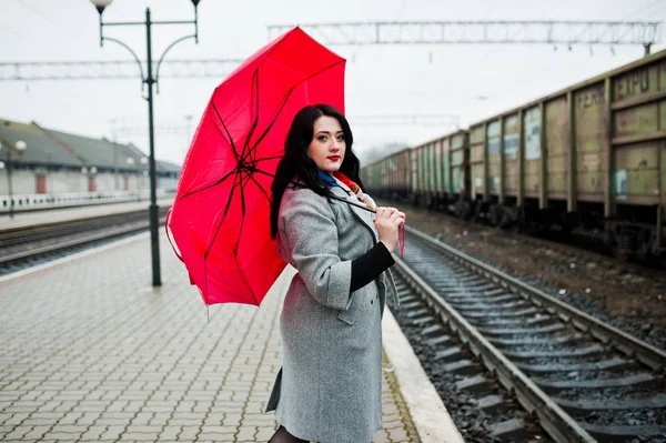 Brunette girl in gray coat with red umbrella in railway station. Royalty Free Stock Photos