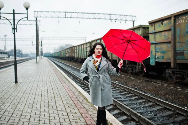 Brunette girl in gray coat with red umbrella in railway station. Stock Picture