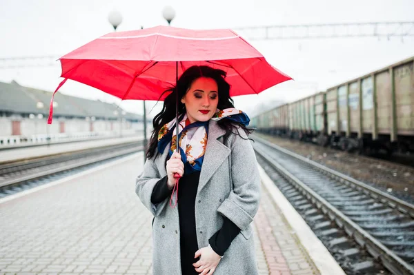 Brunette girl in gray coat with red umbrella in railway station. Royalty Free Stock Photos