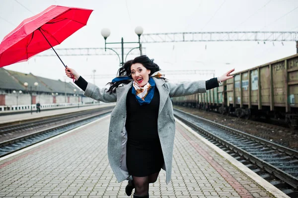 Brunette girl in gray coat with red umbrella in railway station. Stock Photo