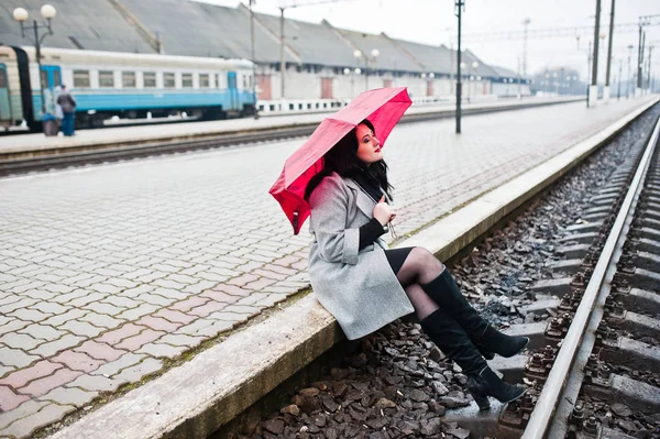 Brunette girl in gray coat with red umbrella in railway station. Stock Image
