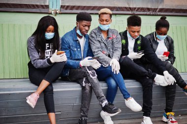 Bir grup Afrikalı genç arkadaş ellerinde telefonlarıyla oturmuş, tıbbi maskeler takıyor. Koronavirüs virüsü virüslerini ve enfeksiyonları önlüyor..