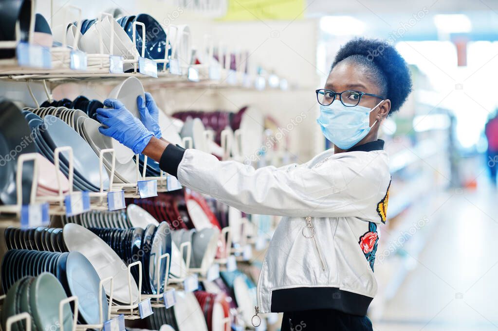 African woman wearing disposable medical mask and gloves shopping in supermarket during coronavirus pandemia outbreak. Black female choose bowl at epidemic time.