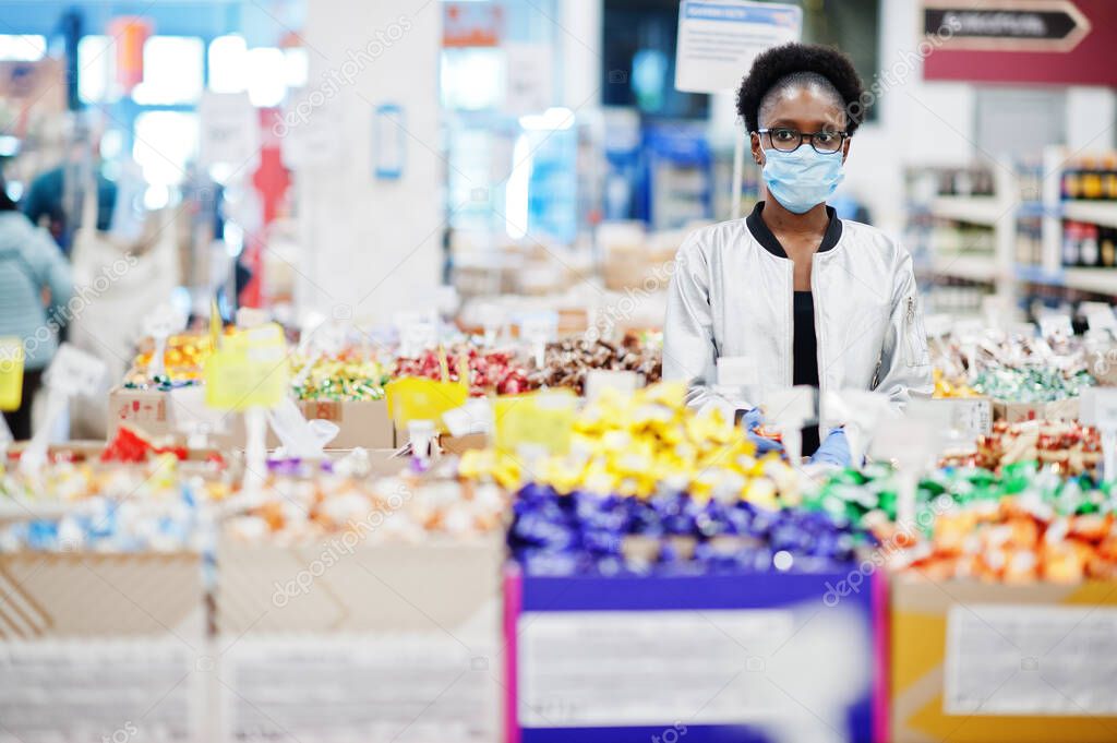 African woman wearing disposable medical mask and gloves shopping in supermarket during coronavirus pandemia outbreak. Black female choose candies at epidemic time.