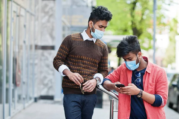Coronavirus covid-19 concept. Two south asian indian man wearing mask for protect from corona virus looking at mobile phone. New normal lifestyle post pandemic in India.