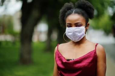 Covid-19, bulaşıcı virüs. Kıvırcık saçlı, kırmızı ipek elbiseli ve tek kullanımlık maskeli bir Afrikalı kadın. Sağlığına önem veriyor ve tehlikeli durumlarda onu koruyor..
