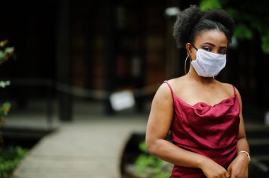Covid-19, bulaşıcı virüs. Kıvırcık saçlı, kırmızı ipek elbiseli ve tek kullanımlık maskeli bir Afrikalı kadın. Sağlığına önem veriyor ve tehlikeli durumlarda onu koruyor..