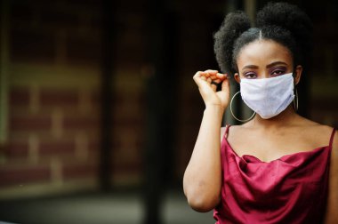 Covid-19, bulaşıcı virüs. Kıvırcık saçlı, kırmızı ipek elbiseli ve tek kullanımlık maskeli bir Afrikalı kadın. Sağlığına önem veriyor ve tehlikeli durumlarda onu koruyor..