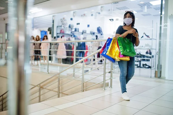 African american woman wearing face protective medical mask for protection from virus disease with shopping bags in mall during coronavirus pandemia.