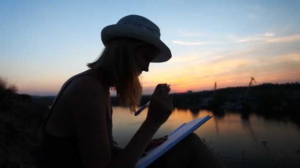 Kreativa personlighet med en anteckningsbok på riverside under solnedgången — Stockvideo