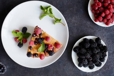 Bir parça yaban mersinli turta, ahududu ve beyaz tabakta tatlı olarak nane şekeri. Arka planda nefis ev yapımı kek parçaları