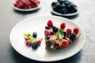 Bir parça yaban mersinli turta, ahududu ve beyaz tabakta tatlı olarak nane şekeri. Arka planda nefis ev yapımı kek parçaları