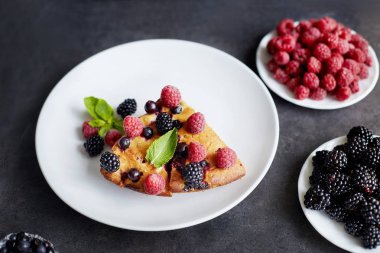 Bir parça yaban mersinli turta, ahududu ve beyaz tabakta tatlı olarak nane şekeri. Arka planda nefis ev yapımı kek parçaları