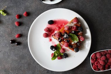 Bir parça yaban mersinli turta, ahududu ve beyaz tabakta tatlı olarak nane şekeri. Arka planda nefis ev yapımı kek parçaları