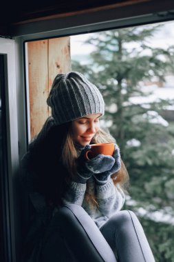 Bir kadının el ele tutuşup kahve içmesi ya da eldivenli kakao içmesi pencere kenarında oturması. Bulanık kar ağacı arka planı. Sabah, rahatlık, kış ve insan konsepti 