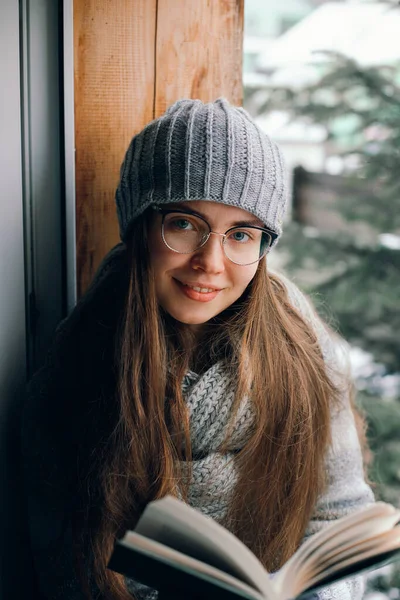 Beautiful woman reading a book in a sweater  sitting home by the window. Blurred winter snow tree background. Morning, coziness, winter and people concept