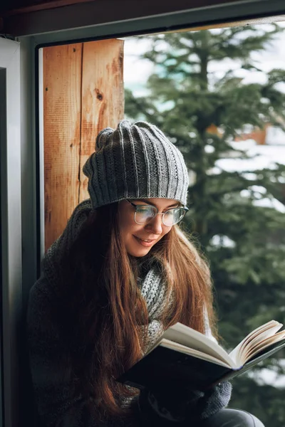 Beautiful woman reading a book in a sweater  sitting home by the window. Blurred winter snow tree background. Morning, coziness, winter and people concept