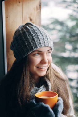Eldivenli bir fincan kahve ya da kakao içen güzel bir kadın pencerede oturuyor. Bulanık kar ağacı arka planı. Sabah, rahatlık ve insan konsepti 
