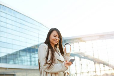  Elinde kahve tutan ve ofisin yakınında telefon kullanan bir iş kadını. Açık havada duran, telefonla gülümseyen güzel bir kadının portresi. Telefon iletişimi. 