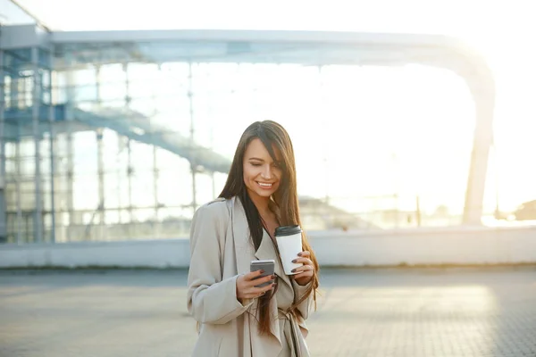 Business woman with coffee and talking on the phone near office. Portrait of beautiful smiling female with phone, standing outdoors. Phone communication.