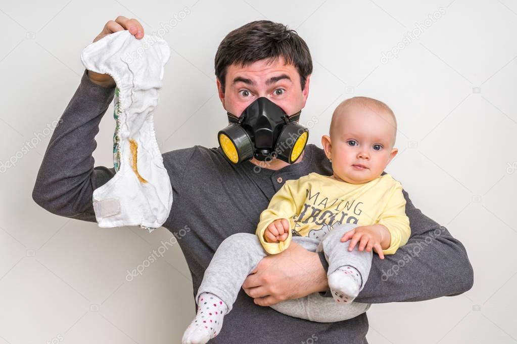 Father with gas mask is holding stinky diaper and little baby