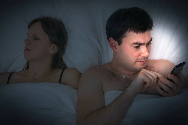 Jovem usando telefone celular, enquanto sua esposa dorme à noite — Fotografia de Stock