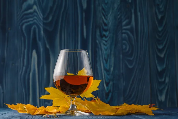 Ποτήρι κονιάκ ή μπράντυ με αποξηραμένα φύλλα φθινοπώρου στο τραπέζι — Φωτογραφία Αρχείου
