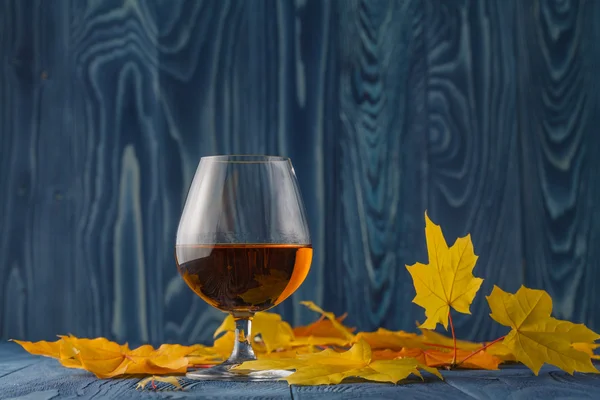 Vidro de conhaque com folhas secas de outono na mesa — Fotografia de Stock