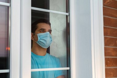 Tıbbi maskeli genç adam pencereden dışarı bakıyor. Koronavirüs salgını. Ev karantinası, Coronavirüs hastalığı yüzünden kendini izole etme, COVID-19. Tıbbi maskeli adam evde kalıyor. Kişisel tecrit.