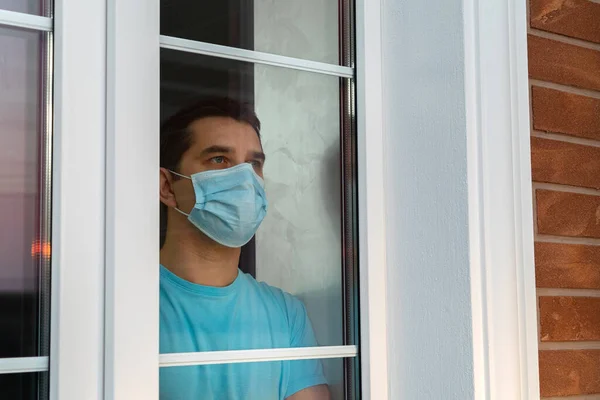 Young man in medical mask is looking out window. Coronavirus pandemic. Home quarantine, self-isolation because of the Coronavirus disease, COVID-19. Man in medical mask stay at home. Self isolation.