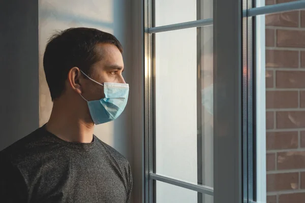 Quarantine self-isolation. Sad young man in a medical mask who looks out the window through the window. Infected man in medical mask on self-isolation looks at the street through the window of a house