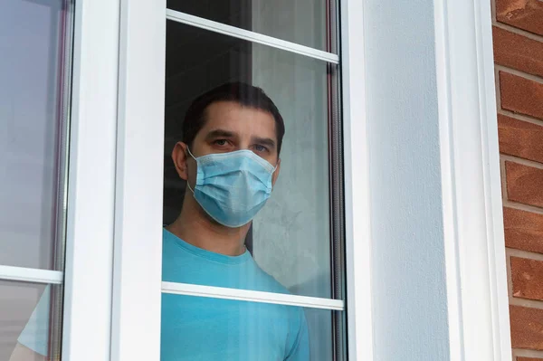 Man in medical mask stay at home. Man in a blue medical mask looks at the camera through a glass window. Self-isolation in the house. Medical mask, protection against coronavirus and other viruses.