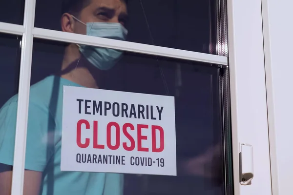 TEMPORARILY CLOSED: sign on a door. Business owner in medical mask puts a CLOSED sign on the front door due to the coronavirus COVID19 pandemic. Small business incurs losses during the coronavirus.