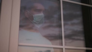 Tıbbi maskeli üzgün adam pencereye bakıyor. 4K