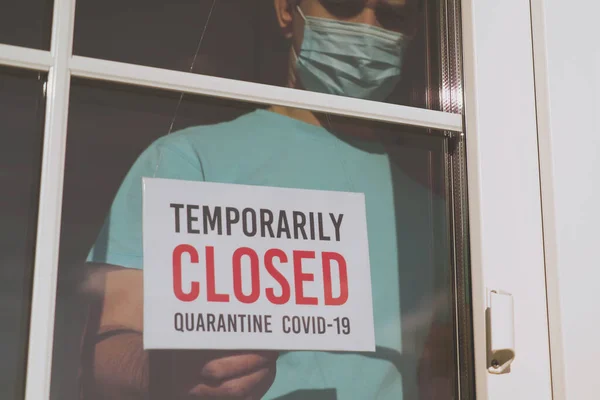 Dükkan penceresindeki küçük işletme tabelası Coronavirus veya COVID19 yüzünden kapanmış. Tıbbi maskeli bir işletme sahibi, Coronavirus COVID19 salgını nedeniyle ön kapıya 