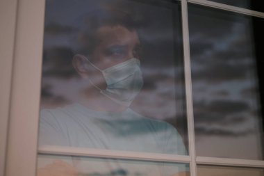 Mesafe için kişisel izolasyon. Karantinanın iptal edilmesini bekliyoruz. Pencereden dışarı bakan, tıbbi maskeli, üzgün bir gencin portresi. Coronavirus COVID-19 karantinasında üzüntü ve özlem.