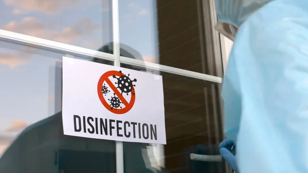 Specialized worker in protective blue overalls stick a DISINFECTION sign and begins preventive disinfection in a public place. Disinfection service, Cleaning and remediation to curb infection.