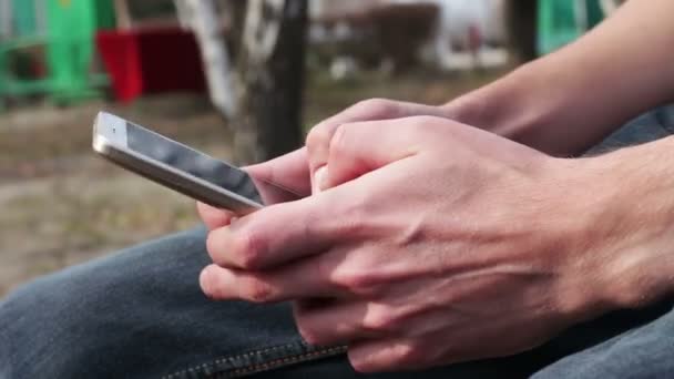 Joven con teléfono blanco se sienta en la calle, en el parque, al aire libre — Vídeo de stock