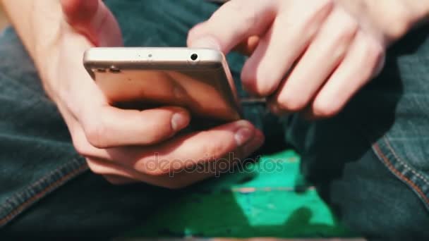 Jovem com telefone branco senta-se na primavera na rua, no parque, ao ar livre . — Vídeo de Stock