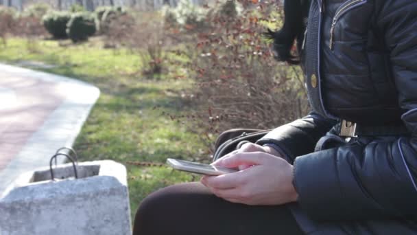 Το κορίτσι εκτυπώνει ένα μήνυμα και κύλιση μέσω των σελίδων σε ένα smartphone σε ένα πάρκο την άνοιξη — Αρχείο Βίντεο