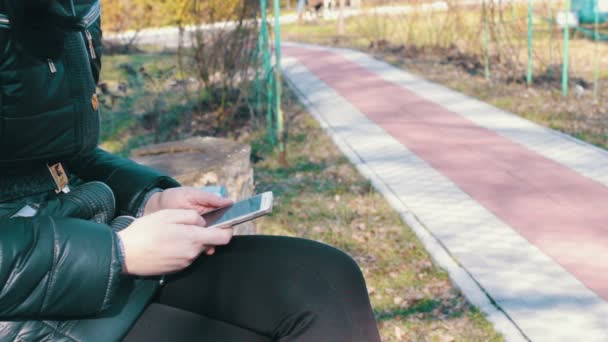 A menina imprime uma mensagem e percorre as páginas em um smartphone em um parque de primavera — Vídeo de Stock