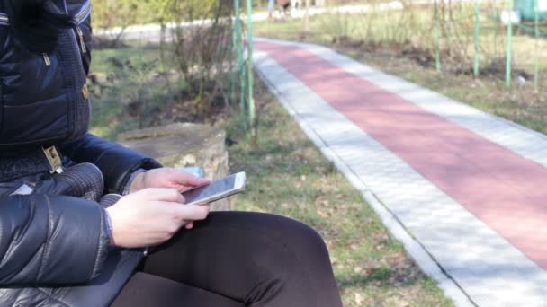 A menina imprime uma mensagem e percorre as páginas em um smartphone em um parque de primavera — Vídeo de Stock