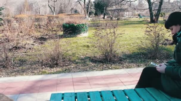 Flickan skriver ett meddelande och bläddrar igenom sidorna på en smartphone i en spring park — Stockvideo